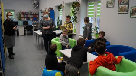 Dzieci siedzą przy stoliku, w tle widać nauczycielkę i bibliotekarkę