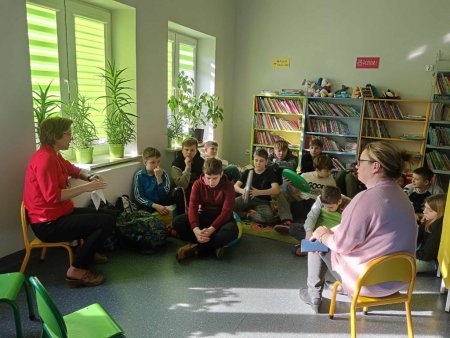 Uczniowie siedzą na dywanie, rozmawiają z bibliotekarką i nauczycielką
