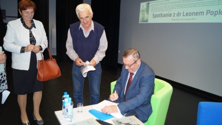 Leon Popek siedzi przy stoliku i podpisuje ksiązki, obok stoją uczestnicy spotkania