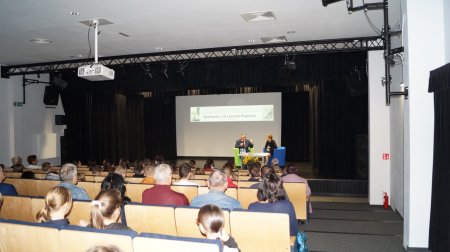 Na sali widowiskowej widać uczestników spotkania z tyłu, na scenie przy stoliku siedzą dr Leon Popek i prowadząca spotkanie