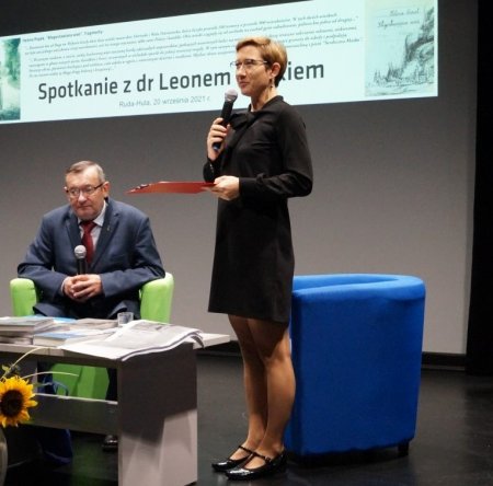 Prowadząca stoi na scenie i mówi do mikrofonu, obok siedzi dr Leon Popek