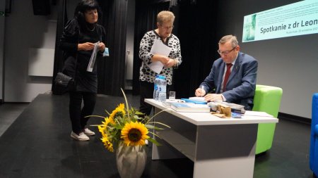 Leon Popek siedzi przy stoliku i podpisuje ksiązki, obok stoją uczestnicy spotkania