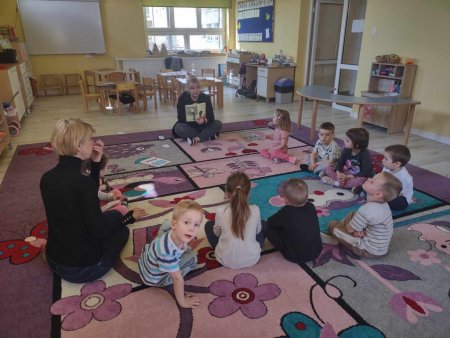 Bibliotekarka siedzi na dywanie i czyta ksiązkę zgromadzonycm wokół dzieciom