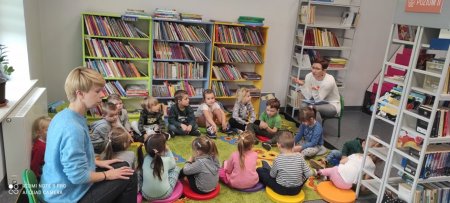 Przedszkolaki siedzą na dywanie, bibliotekarka czyta im bajke, w tle regały z książkami